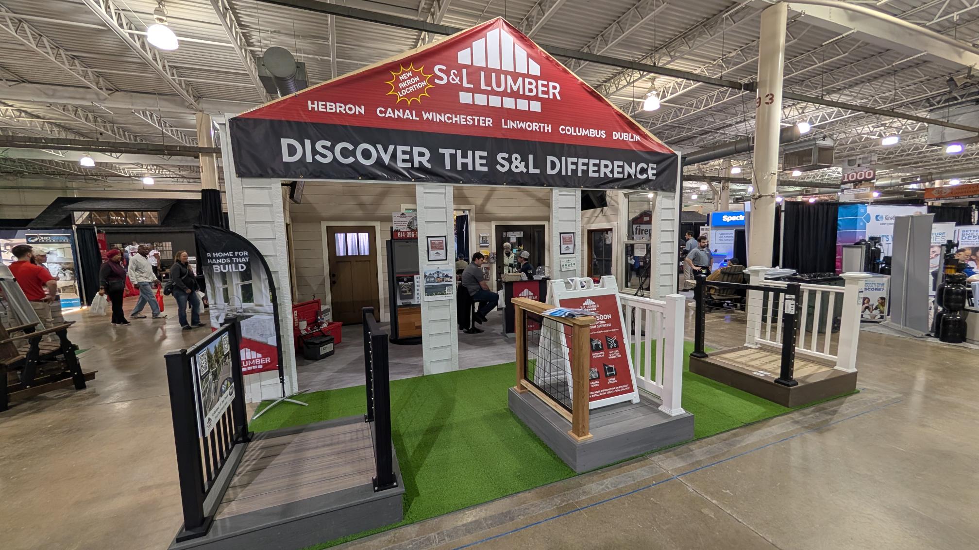 Trade show booth for S&L Lumber with red peaked roof banner, white siding, a small porch, and visitors exploring displays.