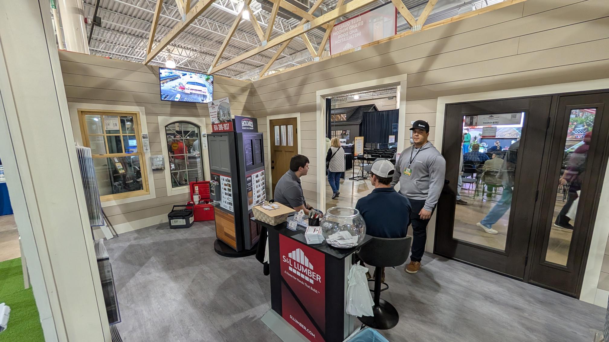 S&L Lumber trade show booth with display panels, sample doors, a glass fishbowl, and staff talking with a visitor.
