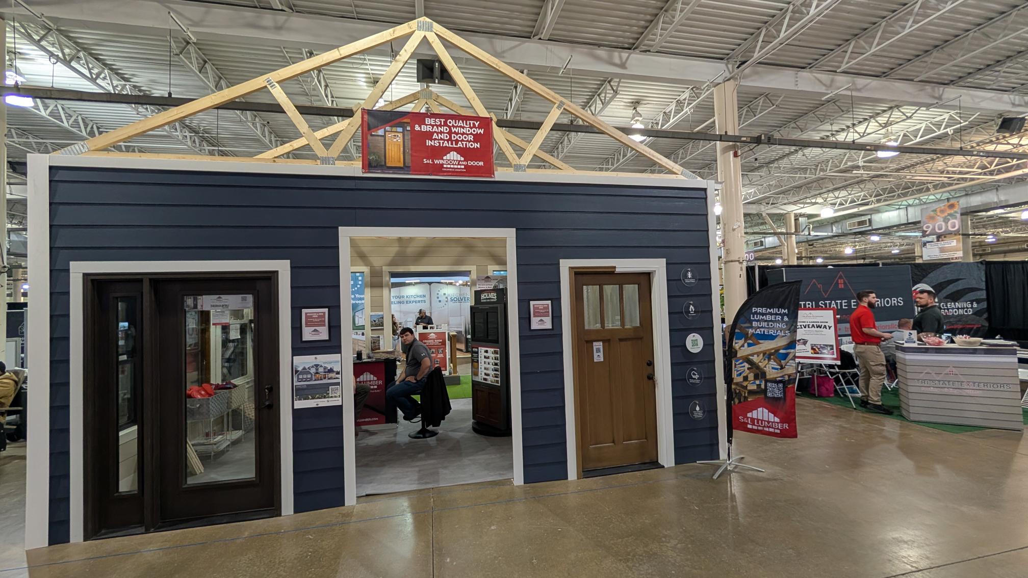 Exhibition booth shaped like a blue house with white trim, featuring doors and wooden roof trusses and a red banner.