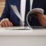 Man dressed in a blue suit flipping through pages on a magazine placed on a desk.