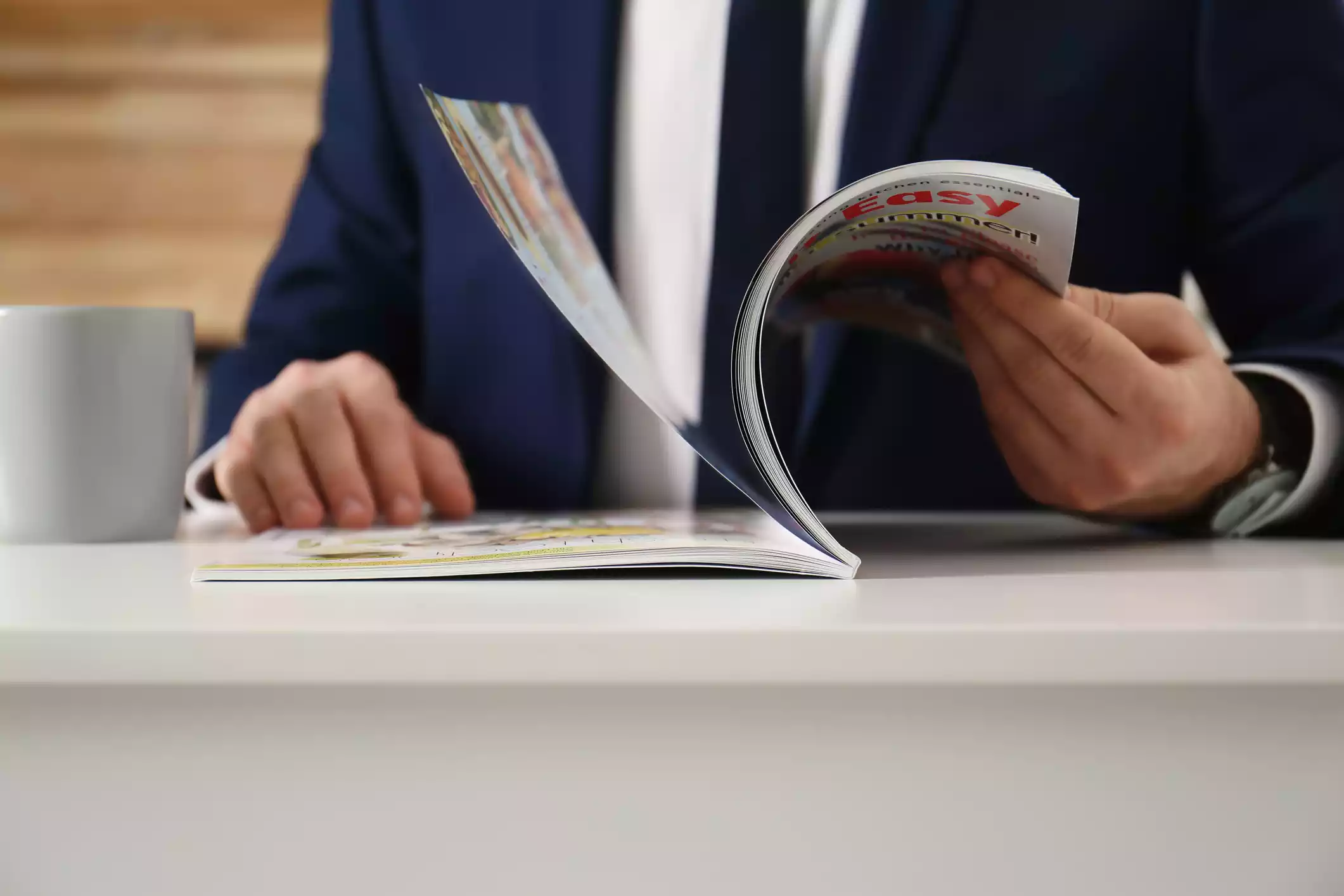 Man dressed in a blue suit flipping through pages on a magazine placed on a desk.